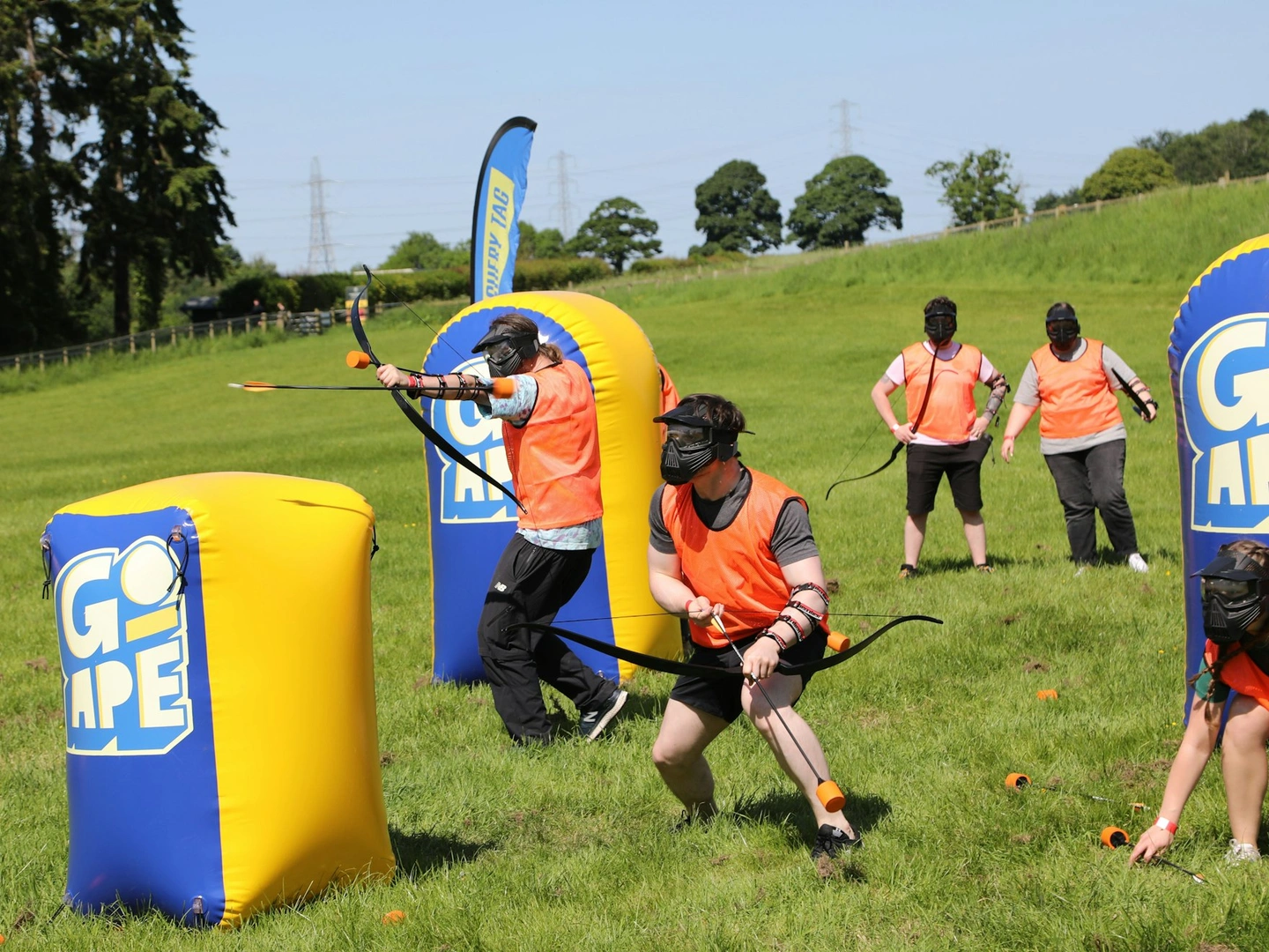 People playing Archery Tag in safety gear using bow and foam-tipped arrows while hiding behind inflatable obstacles.