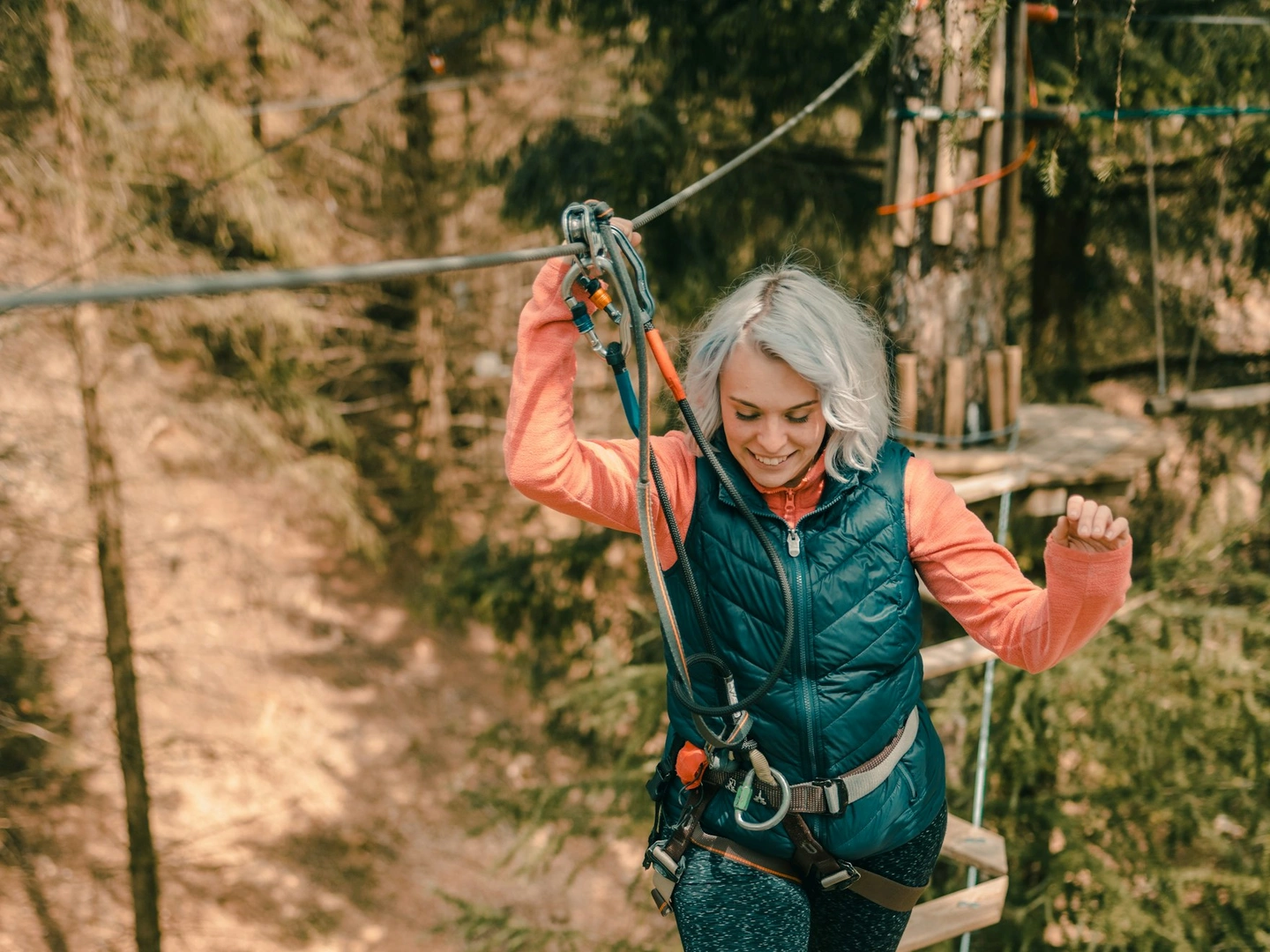 A woman wearing a pink top and gilet smiles as she balances her way over a tree top crossing in the forest