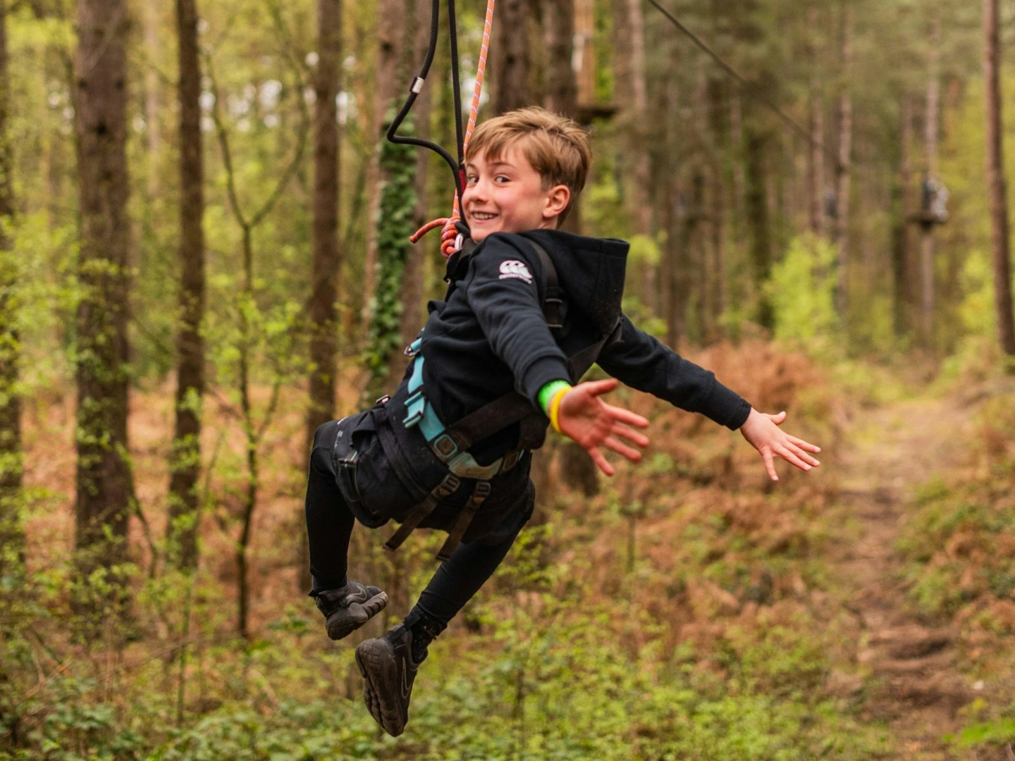 A boy with his arms stretched out and smiling as he rides a zip wire through the forest