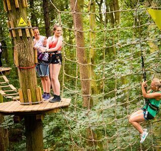 three woman on high ropes course, two stood on platform one swinging into net .