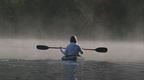 A woman in a kayak on a lake with mist rising from the water and a forest in the background