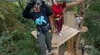 A woman carefully negotiates a high ropes crossing high above the forest floor whilst her colleague watches on from a platform behind